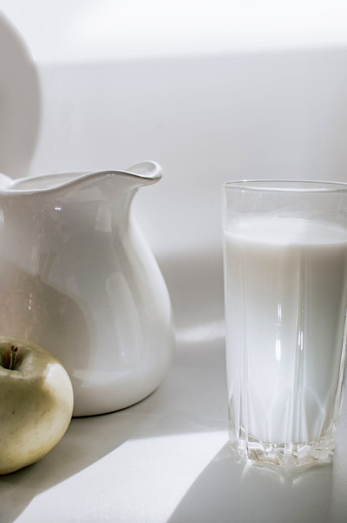 A serene still life image of milk in a glass next to a green apple and ceramic pitcher.